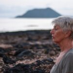 Ältere Frau mit kurzen grauen Haaren sitzt mit geschlossenen Augen an einer felsigen Küste, dahinter Meer und eine Insel